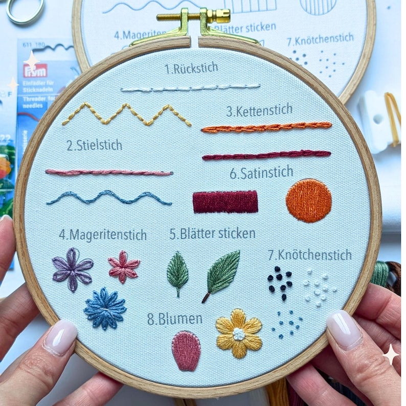 May include: A close-up of an embroidery hoop with various stitches and floral designs. The hoop displays examples of R&uuml;ckstich, Stielstich, and Kn&ouml;tchenstich, along with floral and leaf patterns. The text includes stitch names in German.
