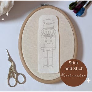 Könnte beinhalten: Ein Stickrahmen mit beigem Stoff und einem Papierschnittmuster eines Nussknackers. Der Text "Stick and Stich Nussknacker" befindet sich auf einem braunen Kreis. Eine kleine Schere und Stickgarne sind ebenfalls zu sehen.