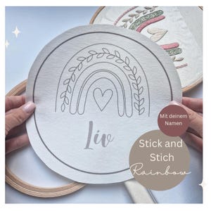 Könnte beinhalten: Ein weißer Stickrahmen mit einer schwarzen Linienzeichnung eines Regenbogens mit Blättern und einem Herzen in der Mitte. Der Name "Liv" steht unter dem Regenbogen. Der Text "Stick and Stitch Rainbow" ist in Braun auf weißem Hintergrund geschrieben.