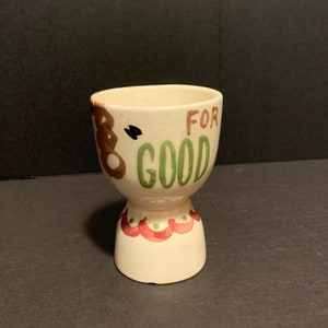 May include: White ceramic egg cup with a green and brown design. The cup has the words "For Good" written on it.