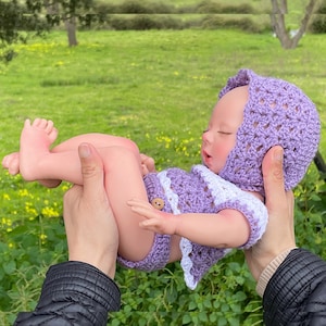 以下が含まれることがあります： 紫と白の編みぐるみベビー人形の衣装と帽子。人形は柔らかなシリコン製で、リアルな顔立ちをしています。