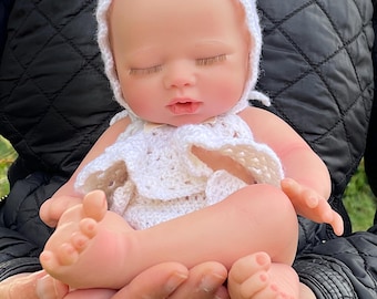 Op de afbeelding: Een realistische babypop met gesloten ogen, die een witte gehaakte muts en een bijpassend outfit draagt. De pop wordt vastgehouden door de handen van een persoon.