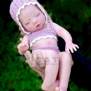 Può includere: Un completo per bambola di crochet rosa con un cappello abbinato. La bambola indossa un top bikini in crochet rosa e bianco e una gonna abbinata. La bambola sta dormendo con gli occhi chiusi.