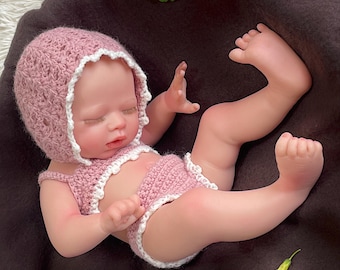 Op de afbeelding: Een levensechte babypop met een roze gehaakte muts en bijpassende tweedelige outfit met witte rand. De pop ligt op een bruin oppervlak, de ogen zijn gesloten. Een gele bloem en groene bladeren zijn op de achtergrond.