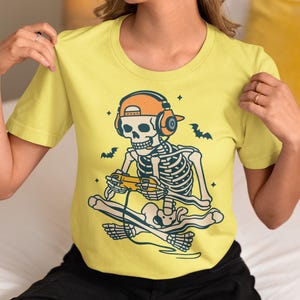 May include: A yellow short-sleeve t-shirt featuring a skeleton gamer design. The skeleton is wearing headphones and a hat, holding a game controller, and sitting on a skateboard. Bats and stars are also included in the design.