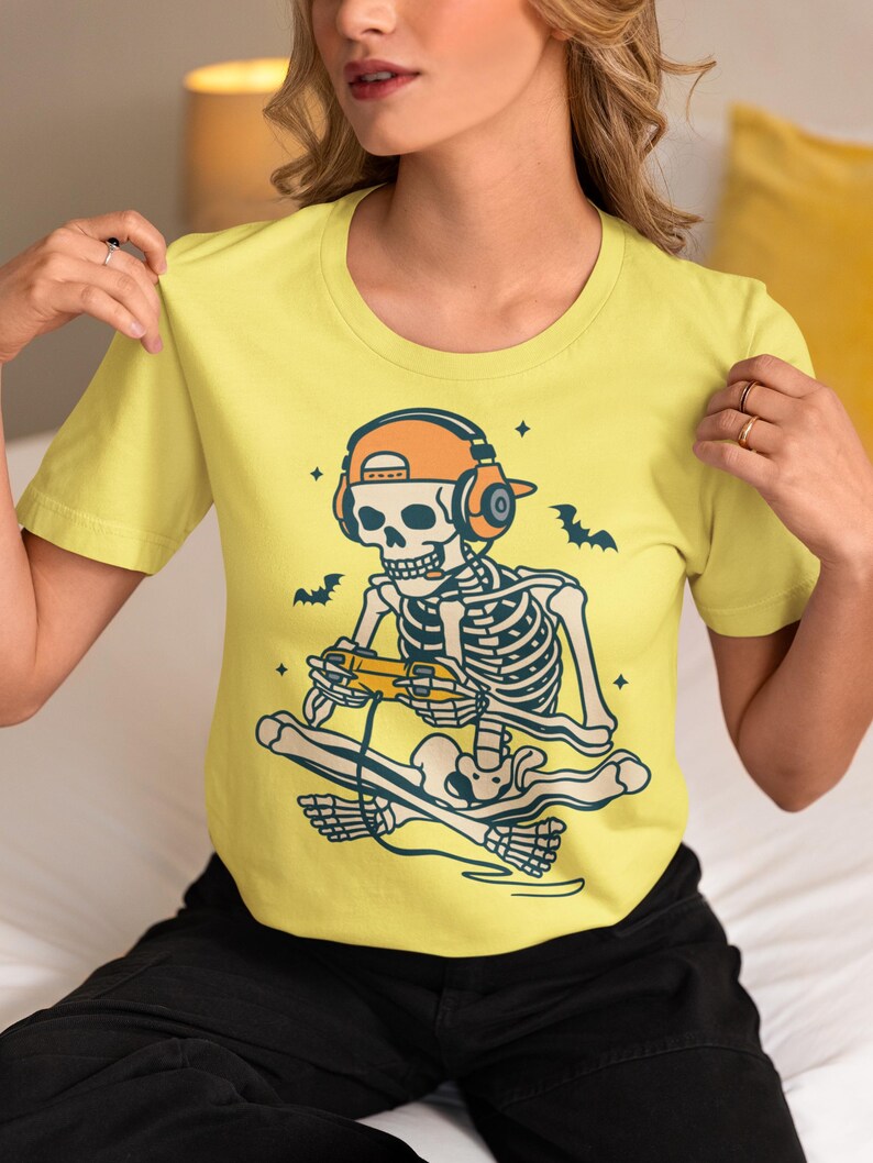 May include: A yellow short-sleeve t-shirt featuring a skeleton gamer design. The skeleton is wearing headphones and a hat, holding a game controller, and sitting on a skateboard. Bats and stars are also included in the design.
