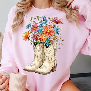 May include: A pale pink sweatshirt with a graphic of cream-coloured cowboy boots filled with colourful flowers. The flowers include orange, pink, blue, and yellow blooms. The boots have decorative stitching and a small heel.