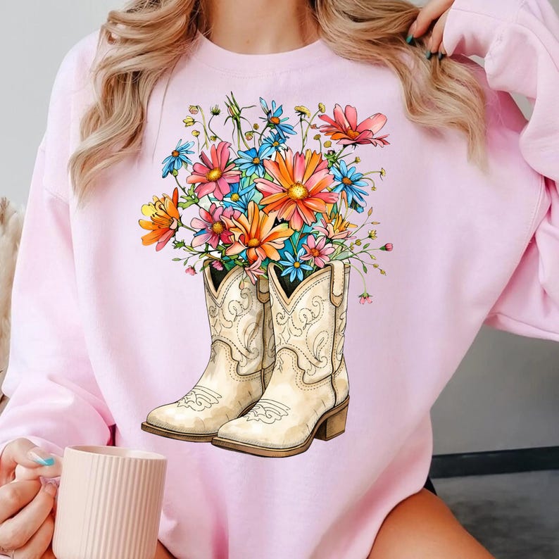 May include: A pale pink sweatshirt with a graphic of cream-coloured cowboy boots filled with colourful flowers. The flowers include orange, pink, blue, and yellow blooms. The boots have decorative stitching and a small heel.