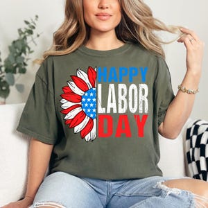 Pode incluir: Camiseta verde-oliva com um design patri&oacute;tico de girassol. As p&eacute;talas do girassol s&atilde;o vermelhas e brancas, com um centro azul cheio de estrelas brancas. As palavras "HAPPY LABOR DAY" s&atilde;o impressas em azul, branco e vermelho.