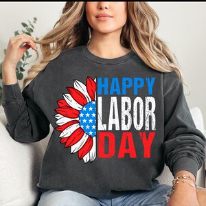 Pode incluir: Camisola cinza escuro com um design patri&oacute;tico de girassol. As p&eacute;talas do girassol s&atilde;o vermelhas, brancas e azuis, com um centro azul contendo estrelas brancas. As palavras "HAPPY LABOR DAY" s&atilde;o impressas em azul, branco e vermelho.
