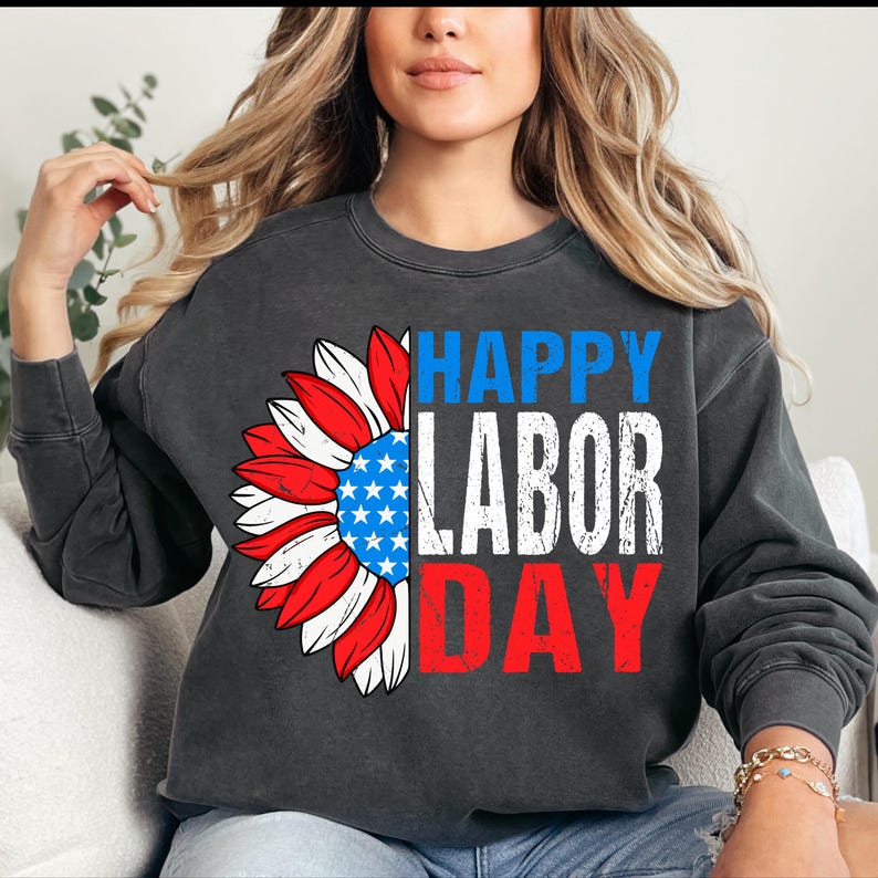 Pode incluir: Camisola cinza escuro com um design patri&oacute;tico de girassol. As p&eacute;talas do girassol s&atilde;o vermelhas, brancas e azuis, com um centro azul contendo estrelas brancas. As palavras "HAPPY LABOR DAY" s&atilde;o impressas em azul, branco e vermelho.