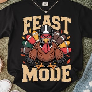 Op de afbeelding: Zwart sweatshirt met een kalkoen die een footballhelm draagt en omringd is door American footballs. De tekst "FEAST MODE" is gedrukt in een beige, blokkerig lettertype. De kalkoen heeft een boze uitdrukking.
