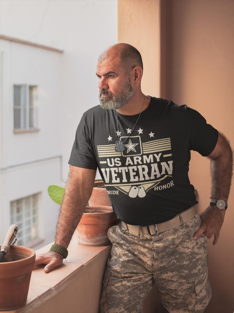 May include: Black t-shirt with "US ARMY VETERAN" text and star graphic. The shirt has a crew neck and short sleeves. The person is wearing camouflage pants and a watch.