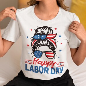 May include: White t-shirt with a graphic design for Labor Day. The design features a woman's face with sunglasses and a headband in red, white, and blue, along with the text "Happy LABOR DAY".