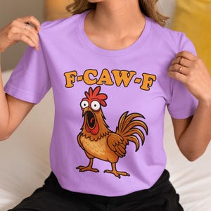 May include: A lilac-colored t-shirt featuring a cartoon rooster with an open beak and the text "F-CAW-F" in orange letters. The rooster is brown with a red comb and wattle.