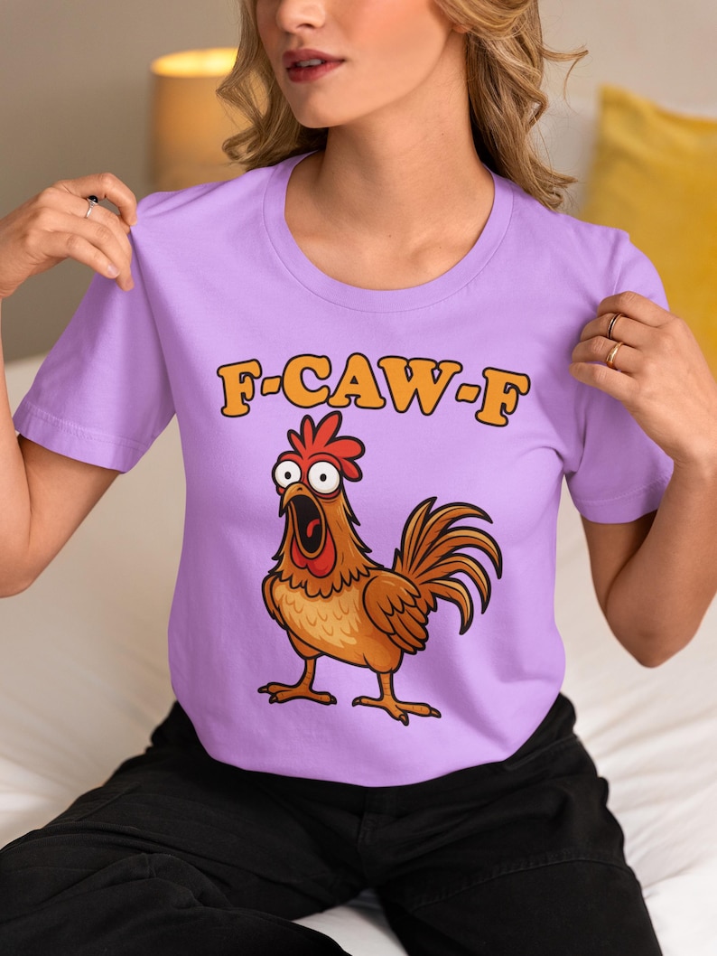 May include: A lilac-colored t-shirt featuring a cartoon rooster with an open beak and the text "F-CAW-F" in orange letters. The rooster is brown with a red comb and wattle.