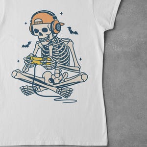 May include: White t-shirt featuring a skeleton gamer with headphones and a controller. The skeleton is cross-legged, with bats and stars around it. The design is in shades of beige, orange, and blue.