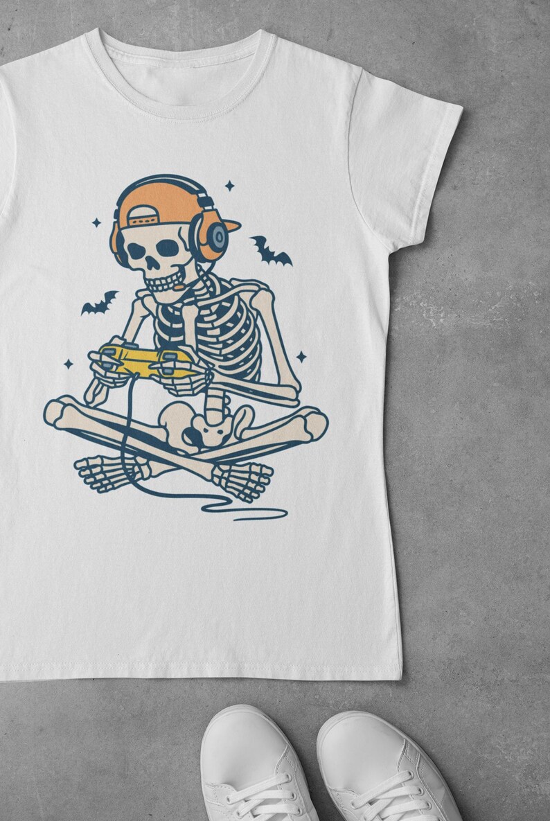 May include: White t-shirt featuring a skeleton gamer with headphones and a controller. The skeleton is cross-legged, with bats and stars around it. The design is in shades of beige, orange, and blue.