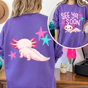 Pu&ograve; includere: Felpa viola con un design di axolotl a cartoni animati e stelle rosa e turchesi. Il retro della felpa presenta un axolotl con branchie rosa e una lunga coda. La parte anteriore mostra il testo "SEE Ya SOON".
