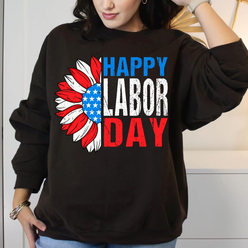Pode incluir: Camisola preta com um desenho de girassol com p&eacute;talas vermelhas, brancas e azuis, e um centro azul com estrelas brancas. O texto "HAPPY LABOR DAY" est&aacute; impresso acima e abaixo do girassol em branco e vermelho.