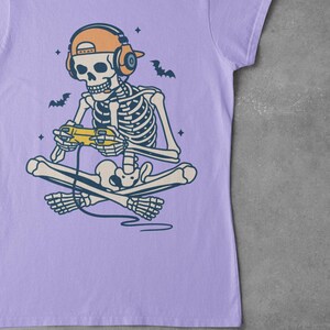 May include: Lilac t-shirt featuring a skeleton gamer with headphones and a controller. The skeleton is cross-legged, with bats and stars around it. The design is a fun, playful take on gaming culture.