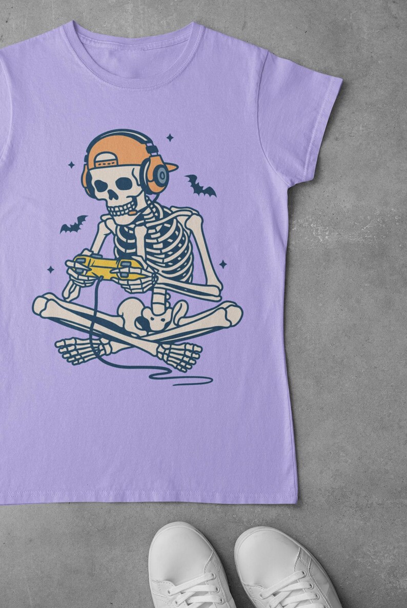 May include: Lilac t-shirt featuring a skeleton gamer with headphones and a controller. The skeleton is cross-legged, with bats and stars around it. The design is a fun, playful take on gaming culture.