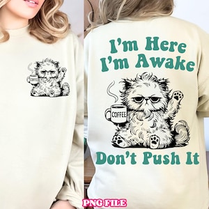 Peut inclure: Sweat-shirt crème avec un motif de chat grincheux tenant une tasse de café. L'avant montre le chat avec une tasse de café, tandis que le dos affiche le texte "I'm Here I'm Awake Don't Push It" en vert d'eau. Un design humoristique pour les amoureux des chats.