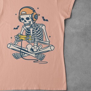 May include: A peach-colored t-shirt featuring a skeleton gamer illustration. The skeleton is seated cross-legged, wearing headphones and a cap, and holding a yellow game controller. Bats and stars are also in the design.