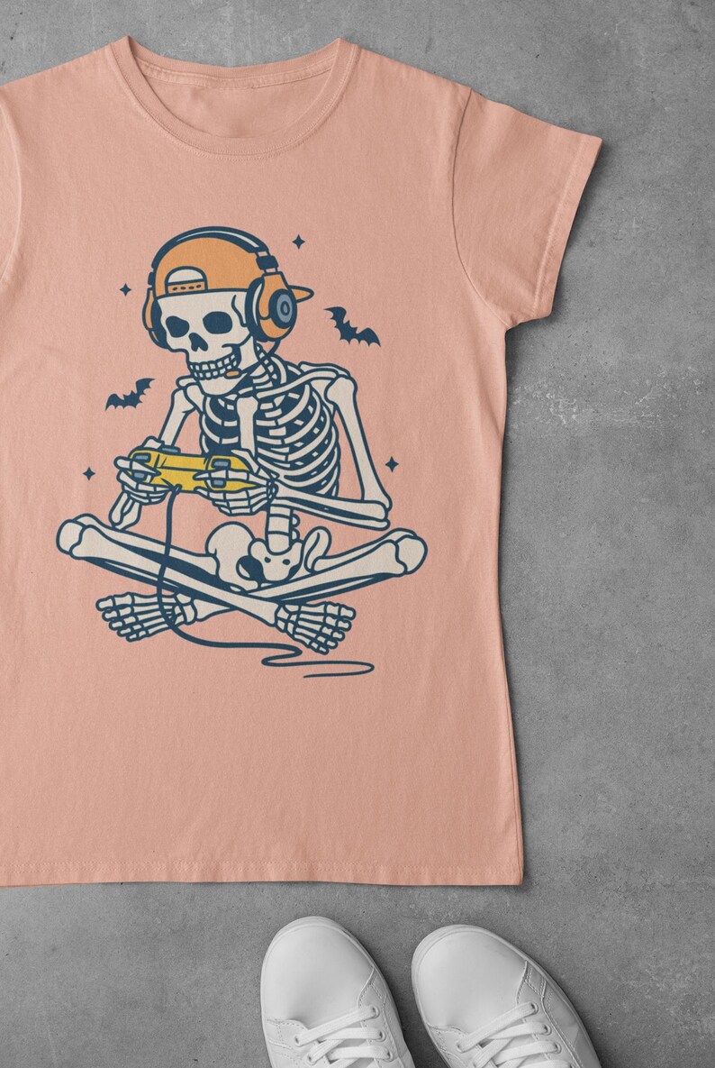 May include: A peach-colored t-shirt featuring a skeleton gamer illustration. The skeleton is seated cross-legged, wearing headphones and a cap, and holding a yellow game controller. Bats and stars are also in the design.