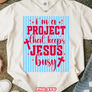 Peut inclure: T-shirt crème avec un motif à rayures bleues et blanches. Le t-shirt affiche le texte "I'm a PROJECT that keeps JESUS busy" en rouge, avec des symboles de croix. Le bas du t-shirt porte le texte "PNG.SVG".