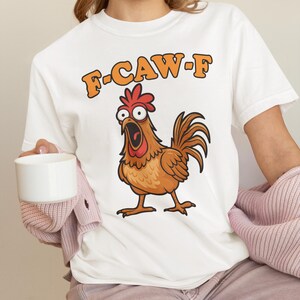May include: White t-shirt featuring a cartoon chicken with an open beak and the text "F-CAW-F" in orange. The chicken is brown with a red comb and wattle. The shirt is a casual, everyday wear item.