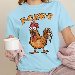 May include: Light blue t-shirt featuring a cartoon chicken with an open beak and the text "F-CAW-F" in orange. The chicken is brown with a red comb and is standing on two feet. The shirt is made of a soft material.