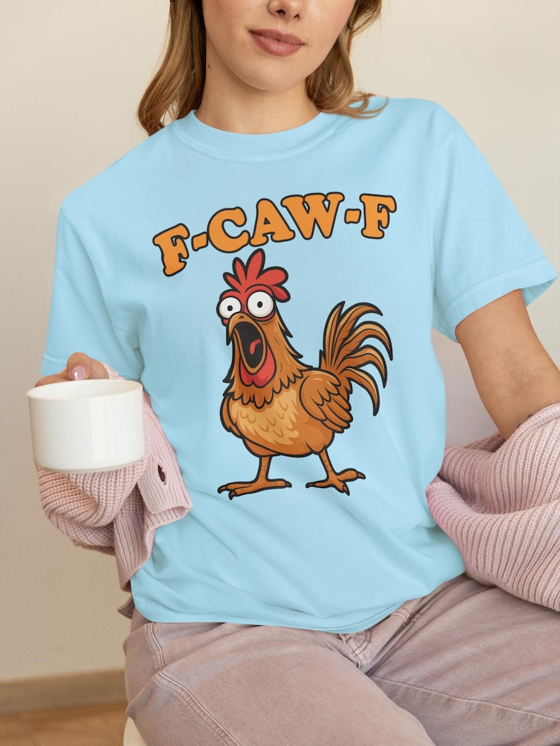 May include: Light blue t-shirt featuring a cartoon chicken with an open beak and the text "F-CAW-F" in orange. The chicken is brown with a red comb and is standing on two feet. The shirt is made of a soft material.