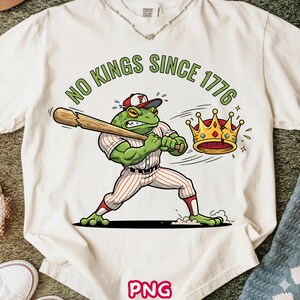 May include: Cream-colored t-shirt with a cartoon frog in a baseball uniform swinging a bat, with the text "NO KINGS SINCE 1776". A gold crown is flying away. The frog is wearing a red and white baseball cap.