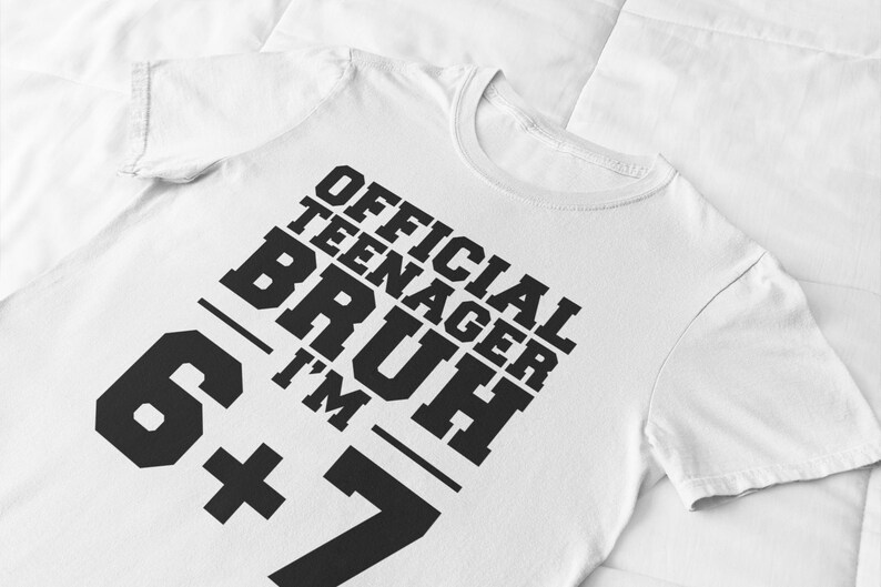 May include: White t-shirt with the text "OFFICIAL TEENAGER BRUH I'M 6+7" in bold black letters. The shirt has a crew neck and short sleeves. The text is arranged diagonally across the front of the shirt.