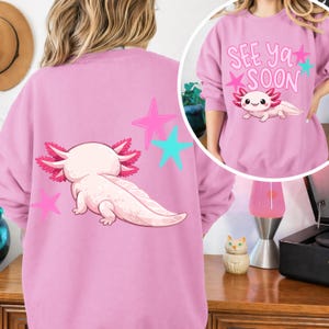 Pu&ograve; includere: Felpa rosa chiaro con un axolotl dei cartoni animati e stelle. L'axolotl &egrave; rosa chiaro con volant rosa. Le stelle sono rosa e turchese. La felpa nell'immagine a riquadro ha il testo "See Ya Soon" e un axolotl.