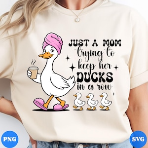 Op de afbeelding: Beige t-shirt met een cartoon eend met een roze handdoek en slippers, een kopje koffie vasthoudend en gevolgd door drie eendjes. De tekst luidt: "JUST A MOM trying to keep her DUCKS in a row."