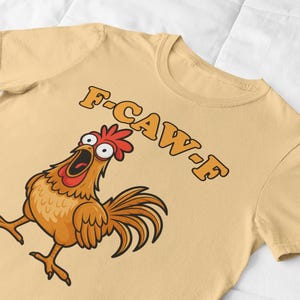 May include: A light tan t-shirt featuring a cartoon chicken with a surprised expression and open beak. The text "F-CAW-F" is printed above the chicken in orange letters. The chicken is brown, orange, and red.