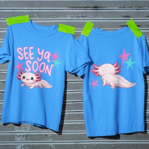 Pu&ograve; includere: Due magliette azzurre con grafiche di axolotl. Una maglietta mostra l'axolotl con la scritta "SEE ya SOON" e stelle. L'altra mostra l'axolotl e le stelle sul retro. Le magliette sono fissate su una superficie metallica.