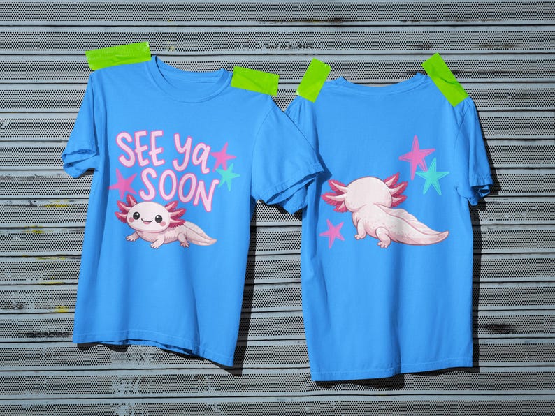Pu&ograve; includere: Due magliette azzurre con grafiche di axolotl. Una maglietta mostra l'axolotl con la scritta "SEE ya SOON" e stelle. L'altra mostra l'axolotl e le stelle sul retro. Le magliette sono fissate su una superficie metallica.