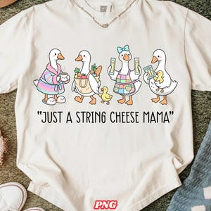 May include: Cream-colored t-shirt featuring a whimsical illustration of four cartoon geese, each carrying different items. The geese are depicted with various accessories and the text "JUST A STRING CHEESE MAMA" is printed below the geese.