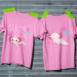 Pu&ograve; includere: Due magliette rosa con un design a cartoni animati di axolotl. Una maglietta mostra il testo "SEE YA SOON" con stelle e un axolotl. L'altra mostra l'axolotl da dietro con stelle. Le magliette sono attaccate a una superficie metallica.
