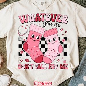 Puede incluir: Camiseta color crema con un gráfico de dos calcetines rosas de dibujos animados con caras sonrientes y decoraciones de corazón. El texto dice "WHATEVER you do DON'T FALL FOR ME". El diseño incluye un estetoscopio y otros gráficos pequeños. El diseño es juguetón y caprichoso.
