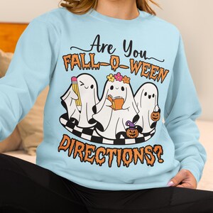 K&ouml;nnte beinhalten: Hellblaues Sweatshirt mit dem Text "Are You FALL-O-WEEN DIRECTIONS?". Die Grafik zeigt drei Cartoon-Geister mit einem Bleistift, einem Buch und einem K&uuml;rbis. Die Geister stehen auf einer schwarz-wei&szlig; karierten Plattform.