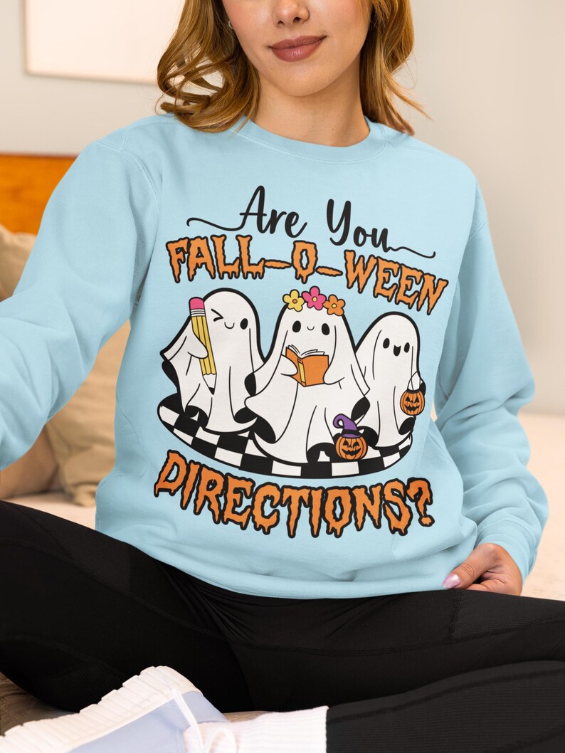 K&ouml;nnte beinhalten: Hellblaues Sweatshirt mit dem Text "Are You FALL-O-WEEN DIRECTIONS?". Die Grafik zeigt drei Cartoon-Geister mit einem Bleistift, einem Buch und einem K&uuml;rbis. Die Geister stehen auf einer schwarz-wei&szlig; karierten Plattform.