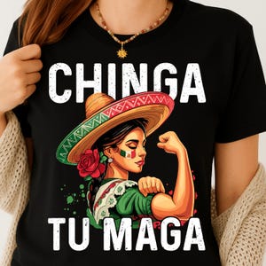 May include: Black t-shirt featuring a graphic of a woman wearing a sombrero, flexing her arm. The text "CHINGA TU MAGA" is printed in white. The woman has the Mexican flag painted on her cheek and is wearing a green dress.