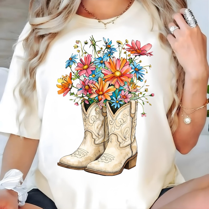 May include: A cream-coloured t-shirt with a graphic of white cowboy boots filled with colourful flowers. The flowers are pink, orange, blue, and yellow. The boots have decorative stitching and a brown heel.