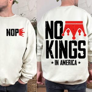 Könnte beinhalten: Cremefarbenes Sweatshirt mit dem Text "NOPE" und einer roten Kronengrafik auf der Vorderseite. Auf der Rückseite des Sweatshirts steht "NO KINGS IN AMERICA" mit einer roten Kronengrafik.