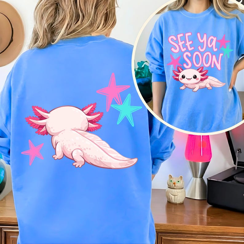 Pu&ograve; includere: Una felpa azzurra con un axolotl dei cartoni animati e la scritta "See Ya Soon". Il design include stelle rosa e turchesi. L'axolotl &egrave; rosa e bianco.