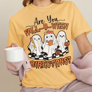 K&ouml;nnte beinhalten: Senfgelbes T-Shirt mit dem Text "Are You Fall-O-Ween Directions?" und einer Grafik von drei Cartoon-Geistern. Ein Geist h&auml;lt einen Bleistift, ein anderer ein Buch und der dritte einen K&uuml;rbis. Die Geister befinden sich auf einem schwarz-wei&szlig;en Schachbrettmuster.